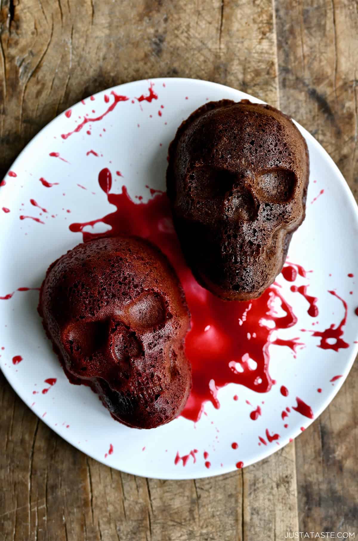 Two mini Halloween skull cakes on a plate, splattered with raspberry sauce.