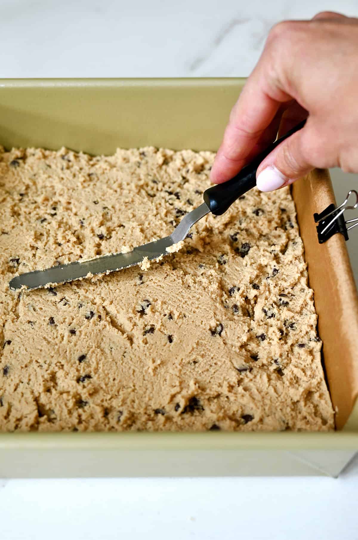 Spreading mini chocolate chip cookie dough into a 9-inch square pan with an offset spatula.