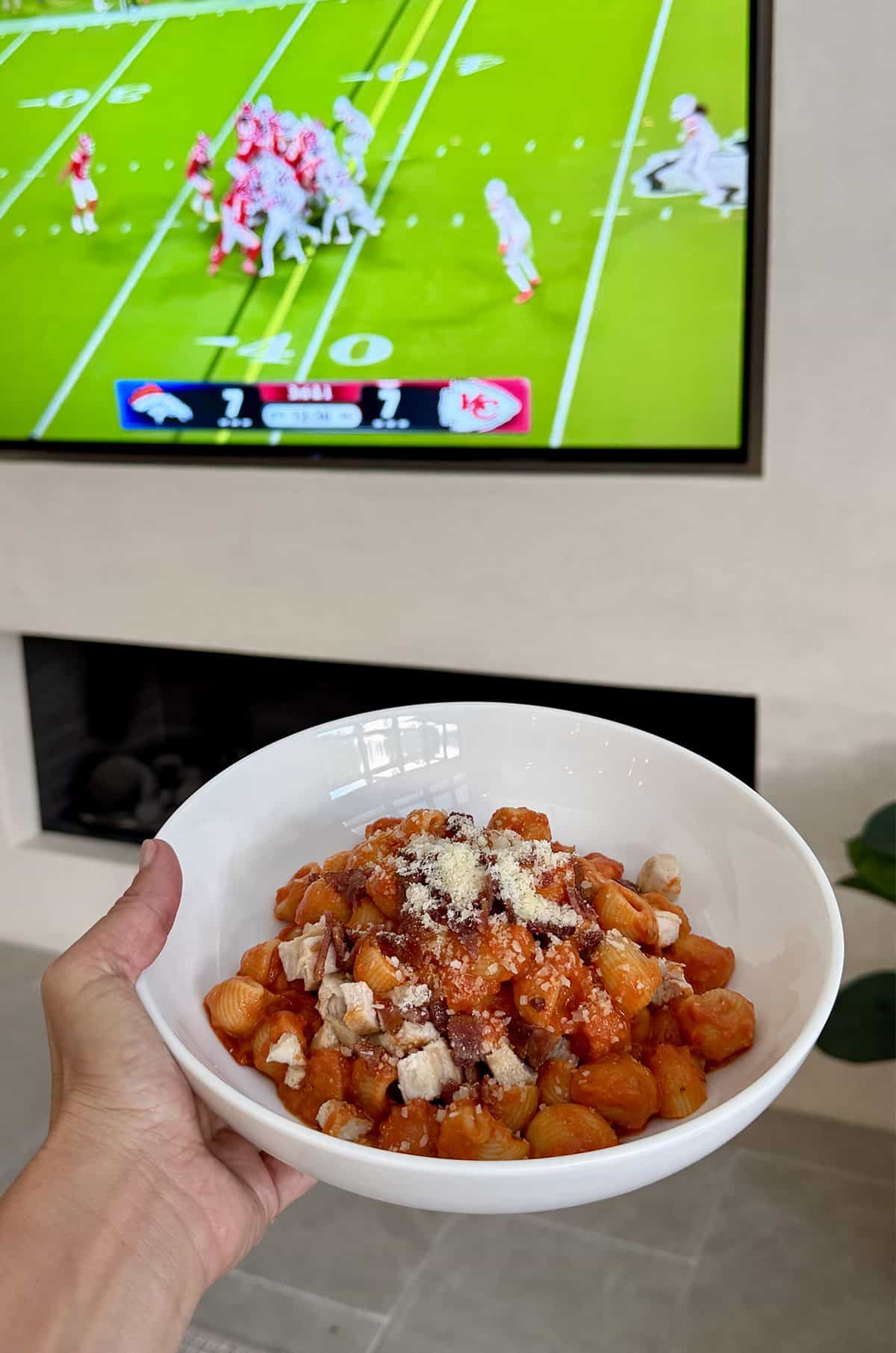 A bowl of vodka pasta with a TV on in the background showing a football game.