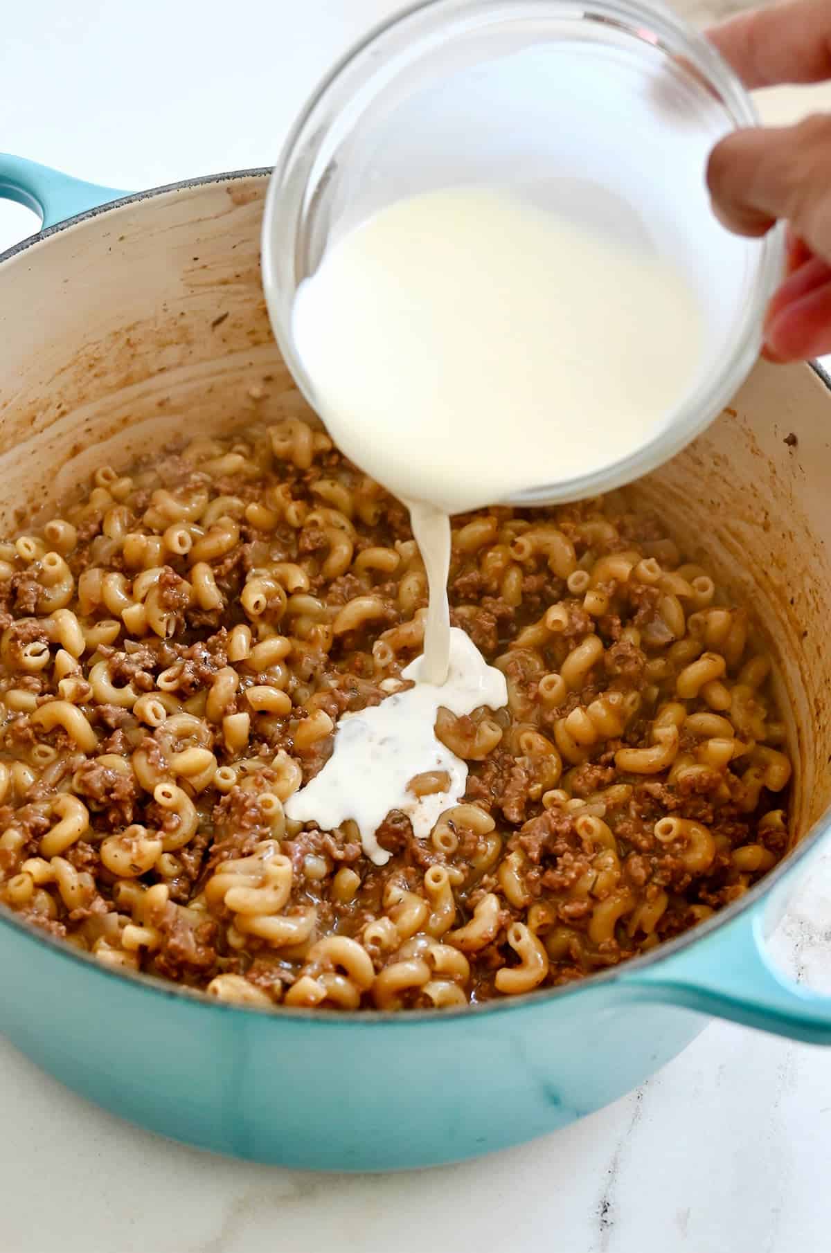 Heavy cream being poured over cooked elbow macaroni and ground beef.